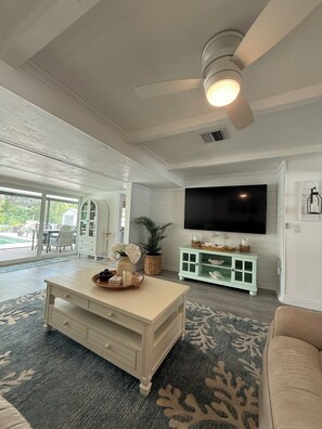 Ein großzügiges Wohnzimmer mit großem Fernseher, Deckenventilator und Blick ins Freie.