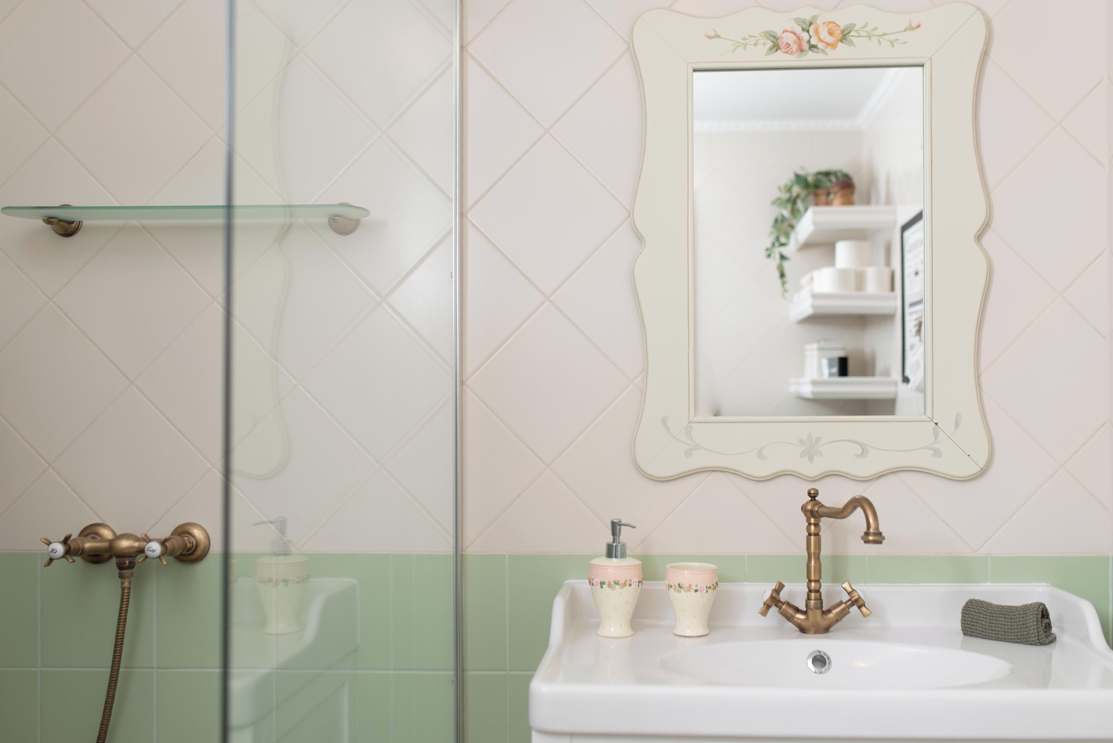 A bathroom with a white sink, a mirror with a floral frame, a shower area, and a glass shelf.