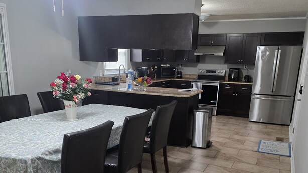 A modern kitchen with a central island, dark cabinetry, stainless steel appliances, and a dining area with a table and chairs.