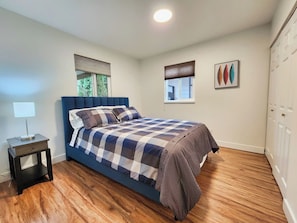 A bedroom with a bed, bedside table, lamp, windows with blinds, and a door.