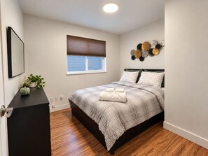 Un dormitorio con cama, ventana con persianas, un adorno en la pared y una pequeña mesa con una planta.