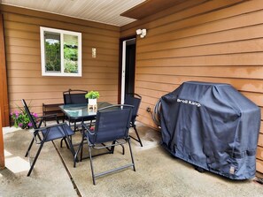 Un patio exterior con mesa y sillas, una parrilla cubierta y una ventana con vistas a zonas verdes.