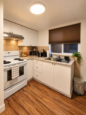 Une cuisine compacte avec des meubles blancs, un four encastré, une cuisinière et une fenêtre avec des stores.