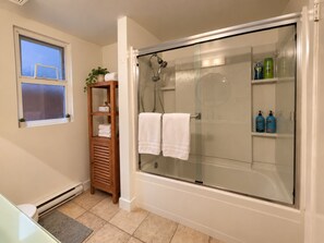 Un baño con ducha envuelta en vidrio, un estante de madera con toallas y una ventana con persianas.