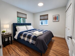 Une chambre à coucher avec un lit, une table de chevet, une lampe, une fenêtre avec des stores et une porte.