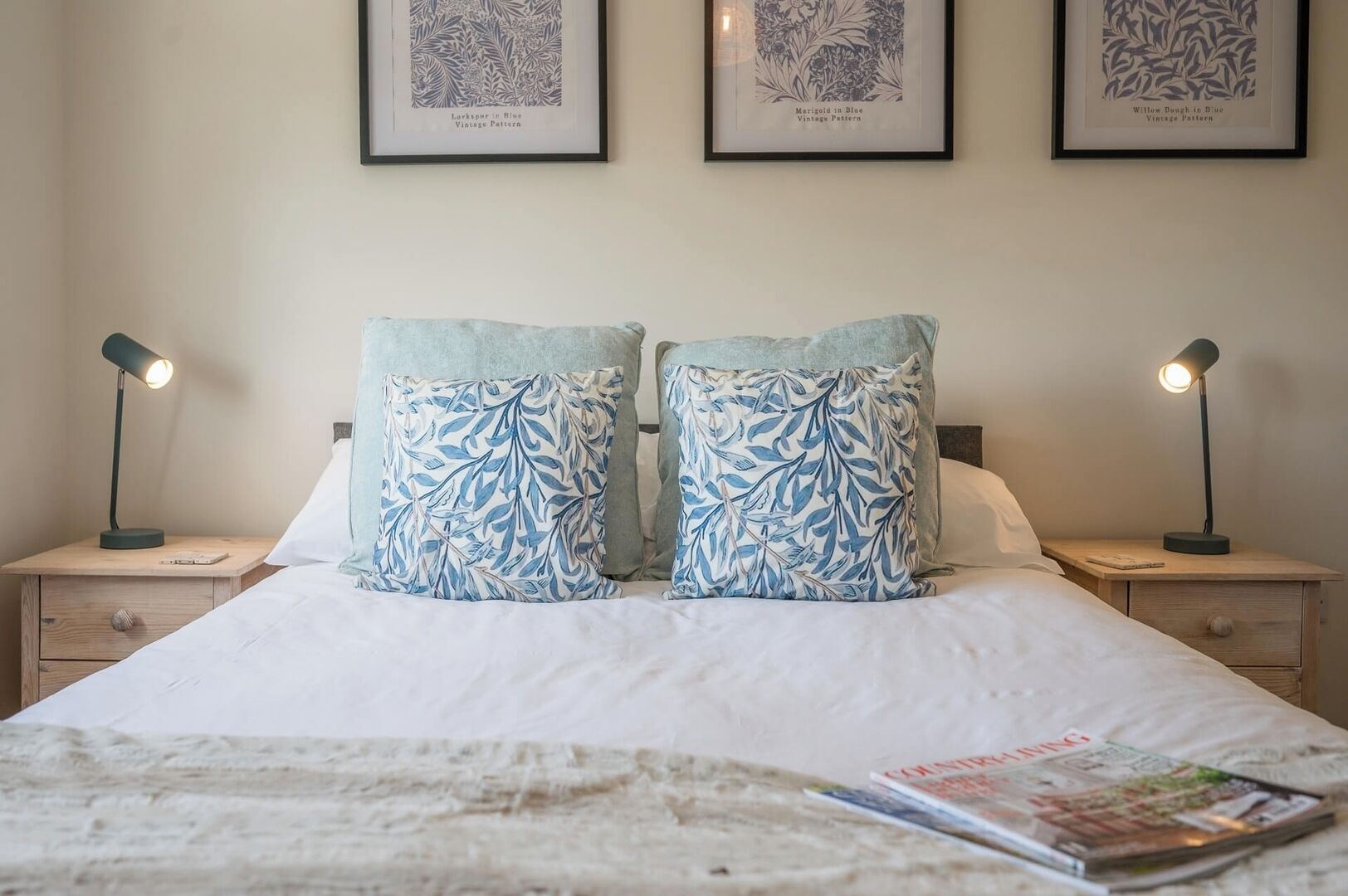 Una cama bien tendida con dos almohadas iguales, una lámpara de noche a cada lado y un patrón antiguo enmarcados en la pared.