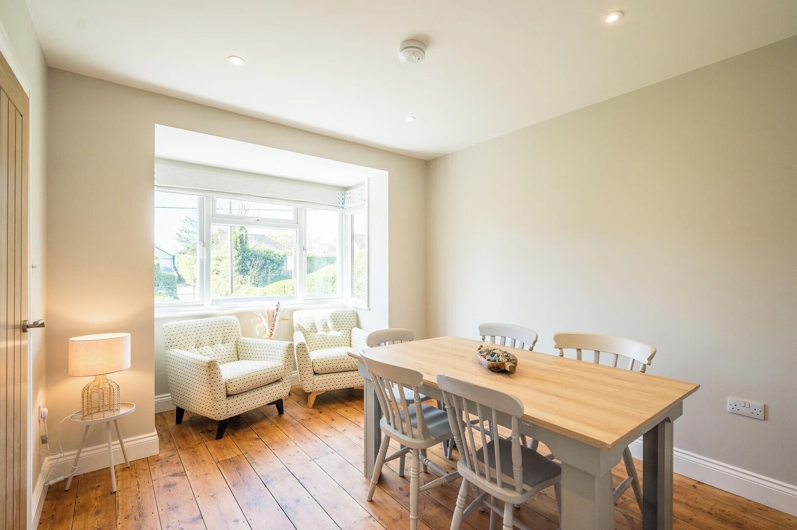 A dining area with a wooden table and chairs, a window with a view of greenery, and a lamp on a small table.