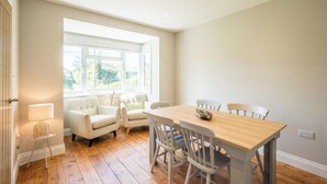 A dining area with a wooden table and chairs, a window with a view of greenery, and a lamp on a small table. - 105 Saxmundham Road, Aldeburgh (Aldeburgh)
