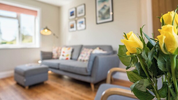 A living room with a grey sofa, a small ottoman, and a vase of yellow flowers. - 105 Saxmundham Road, Aldeburgh (Aldeburgh)