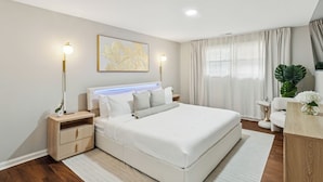 A modern bedroom with a large bed, bedside tables, a nightstand, a headboard with integrated lighting, a large window with curtains, and a potted plant. - 8 BR • Theater • Game Room • Fire Pit • Sleeps 15 (Birmingham)
