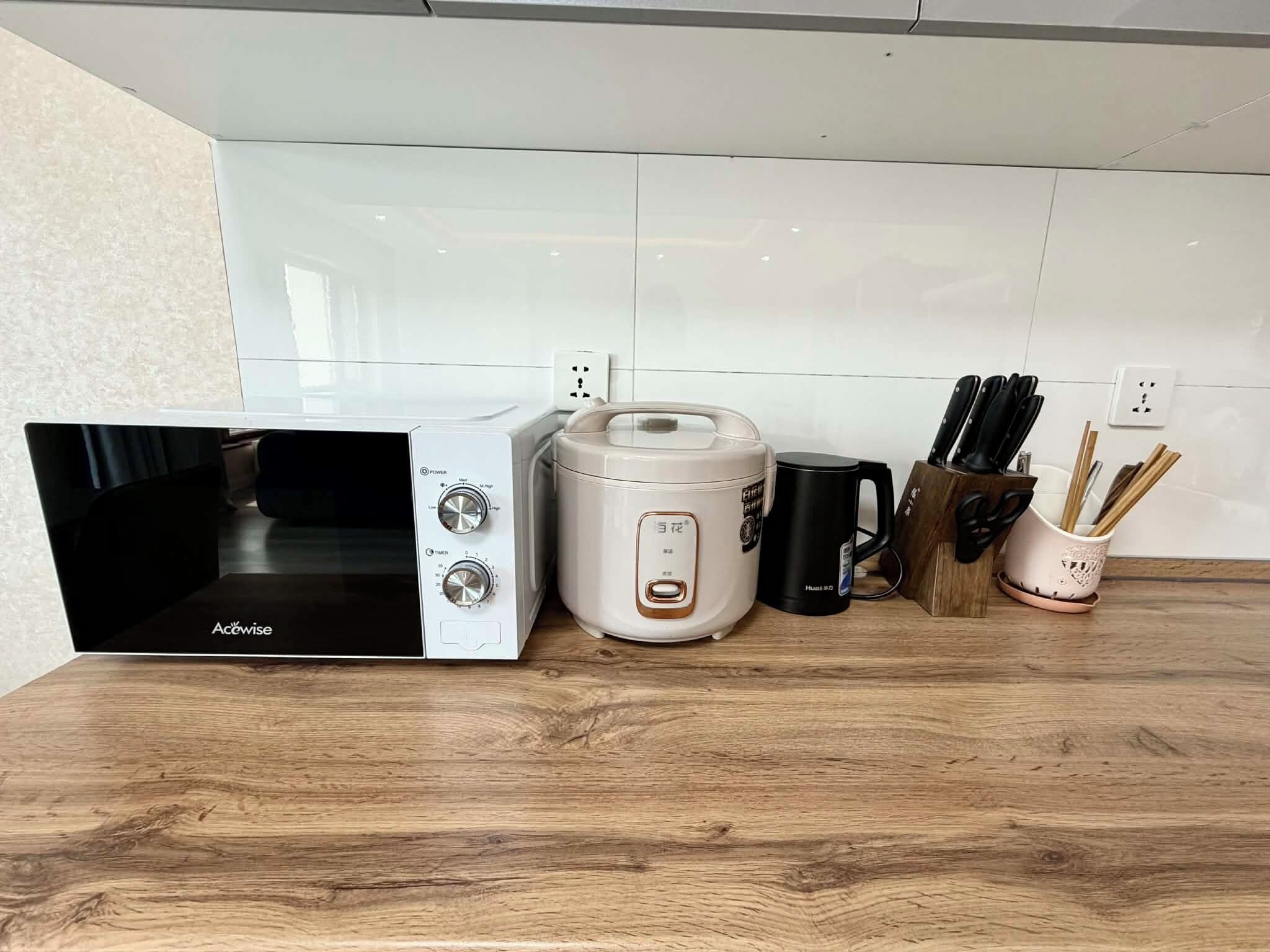 Una cocina con microondas, cocedora de arroz, taza negra y bloque de cuchillos.