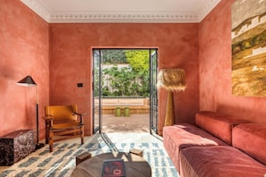 Une pièce avec un mur rose, une chaise en bois, un canapé rouge, un lampadaire et une vue sur la verdure à travers une porte vitrée.