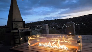 Un barbecue doté d’une enceinte en verre, d’une cheminée en pierre et illuminé de guirlandes lumineuses, le tout sur fond d’arbres et de collines.