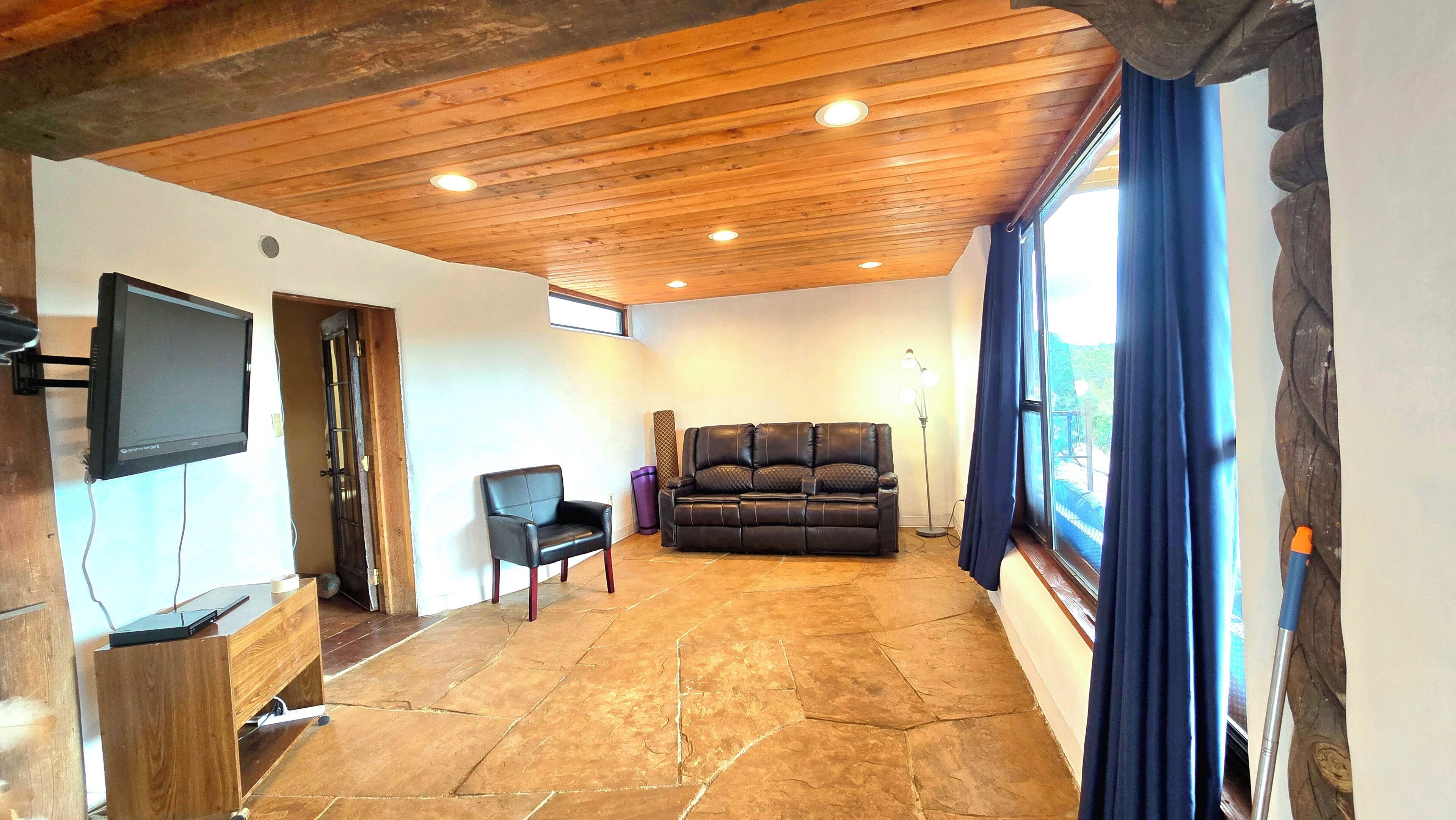 A living room with a stone floor, wooden ceiling, a black leather sofa, a TV mounted on the wall, and a window with curtains.