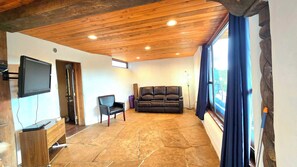 A living room with a stone floor, wooden ceiling, a black leather sofa, a TV mounted on the wall, and a window with curtains.