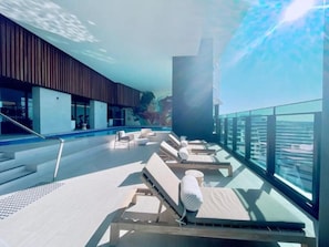 A rooftop pool area with sun loungers, a glass balustrade, and a view of the city skyline. - Luxury High-Rise Stay with Panoramic GC Views (Broadbeach)