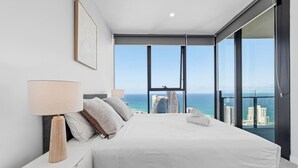 A modern bedroom with a large bed, a nightstand, a lamp, and a view of the ocean and city skyline. - Luxury High-Rise Stay with Panoramic GC Views (Broadbeach)