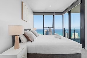 A modern bedroom with a large bed, a nightstand, a lamp, and a view of the ocean and city skyline. - Luxury High-Rise Stay with Panoramic GC Views (Broadbeach)