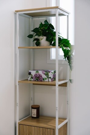 Una estantería blanca con base de madera, una planta en maceta en el estante superior, una caja floral en el estante central y una vela en el estante inferior.