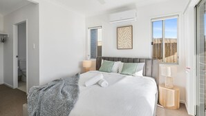 Un dormitorio con cama, ventilador de techo, unidad de aire acondicionado, ventana y puerta que da a un baño.