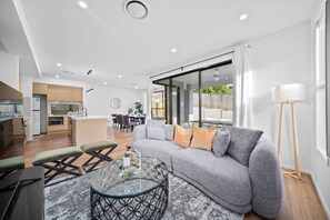 A modern living room with a grey sofa, a glass coffee table, and a dining area in the background. - Luxury New Home| Prime Location|Easy City Access (Mount Gravatt East)
