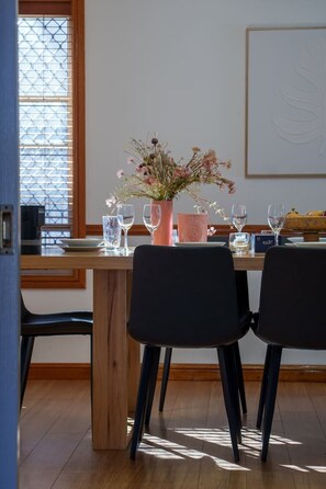 Een eetruimte met een houten tafel, twee zwarte stoelen, een vaas met bloemen en een raam met zonwering.