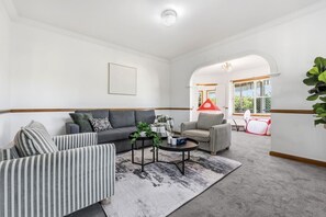 Een ruime woonkamer met een grijze bank, gestreepte fauteuils, een salontafel en een rode, tentachtige constructie op de achtergrond.