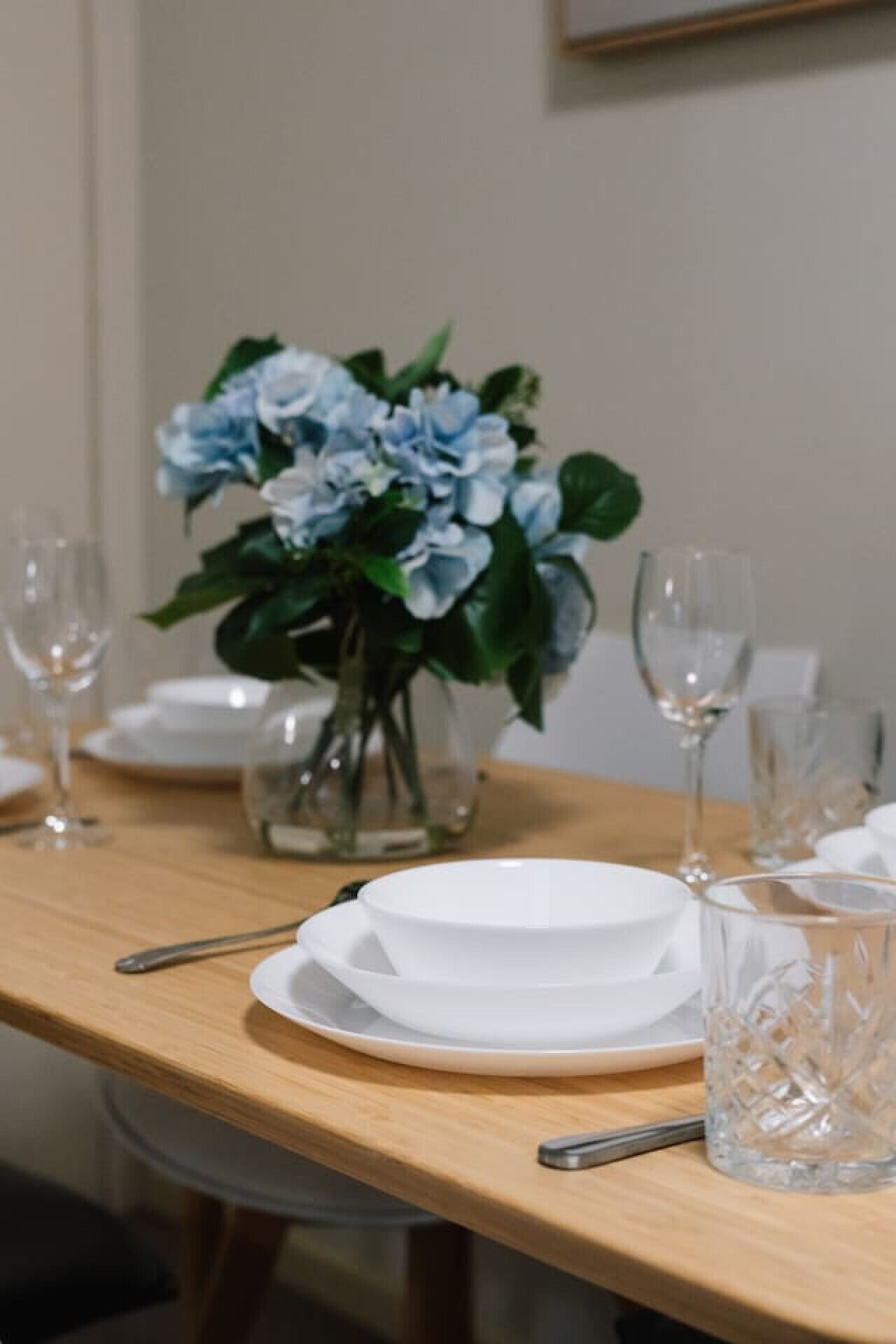 Een eettafel met witte borden, een helder glas en een vaas met blauwe bloemen.