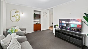 A living room with a grey sofa, a TV on a black stand, a wooden cabinet, and a wall-mounted clock. - Affordable Stay | Private & Convenient |Near Shops (Jindalee)