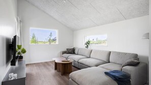 Ein modernes Wohnzimmer mit einem Ecksofa, einem Couchtisch und einem Flachbildfernseher. Zwei Fenster bieten Ausblick ins Freie.