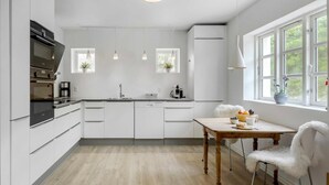 A modern kitchen with white cabinetry, a built-in oven, a coffee machine, and a dining area with a table and chairs. - VillaVilla 953 - Fjaltring-Trans, Vestjylland (Lemvig)