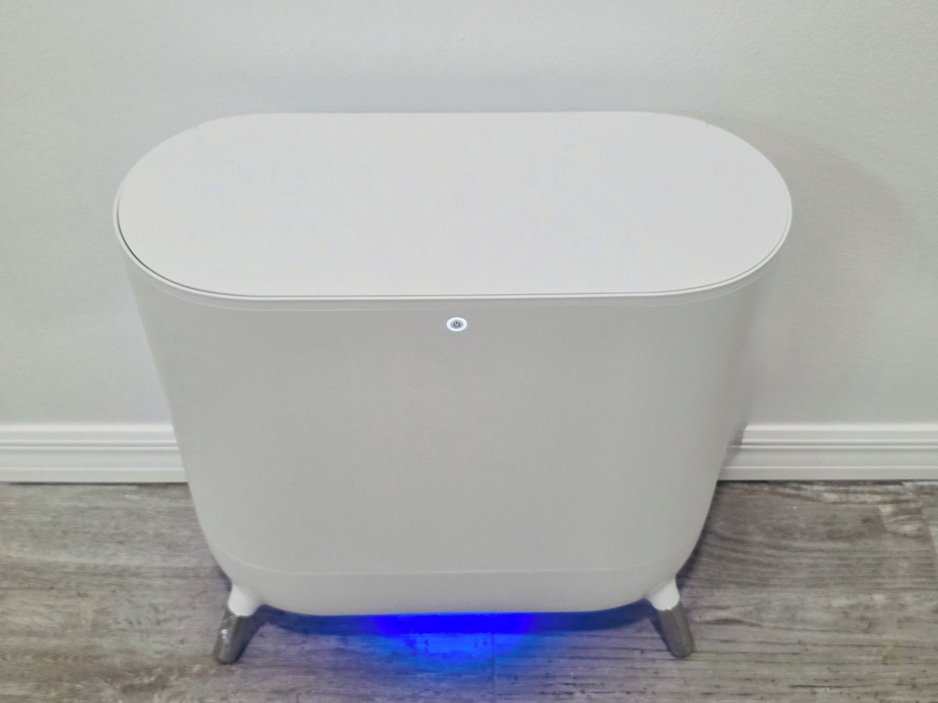 A white, standalone trash bin with a circular button on top, placed on a wooden floor against a plain wall.