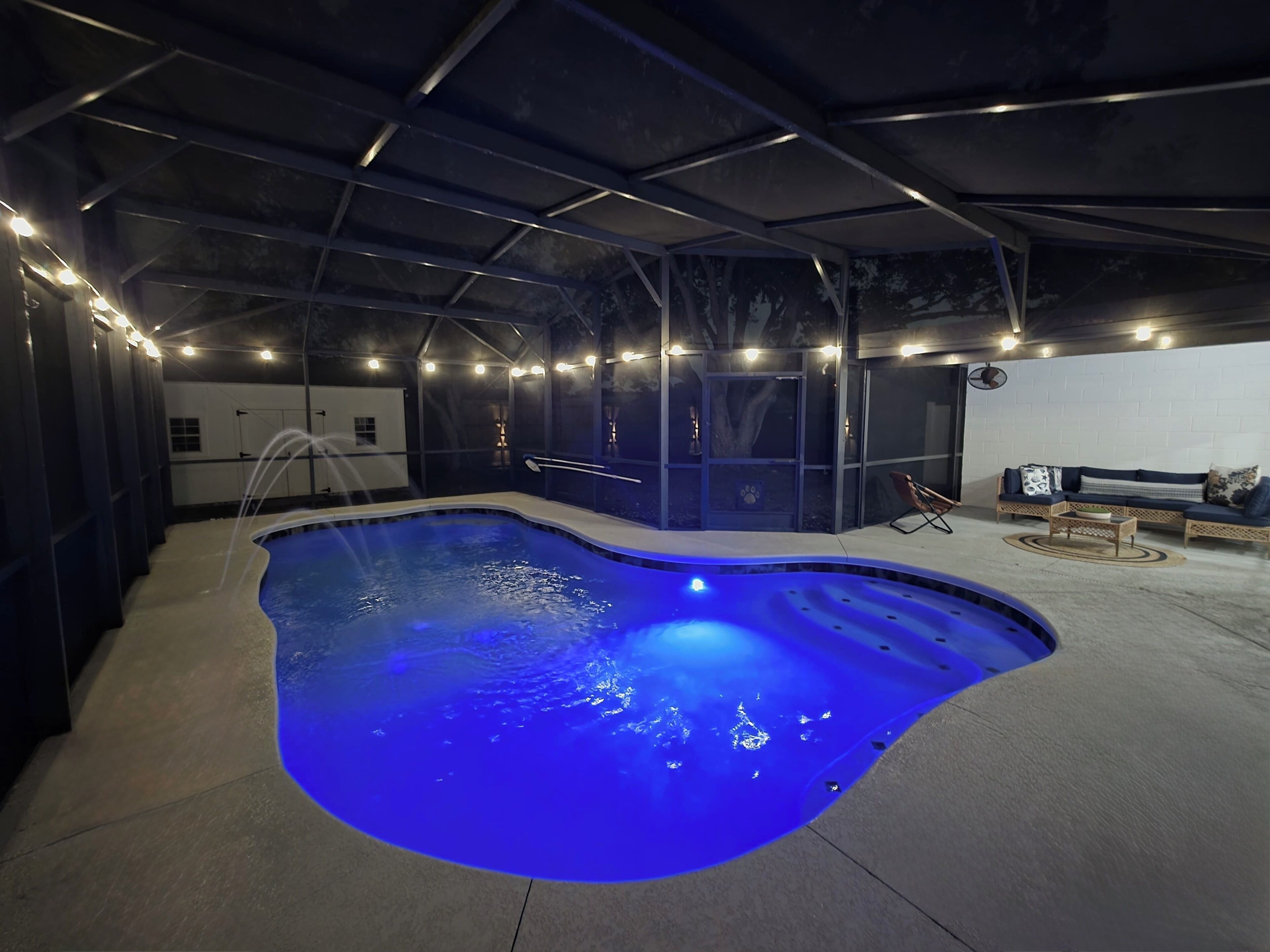 A swimming pool with a blue light, a lounge area with a sofa and chairs, and a white brick wall.