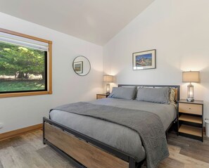 Ein Schlafzimmer mit einem Bett, zwei Nachttischen, einem Fenster mit Blick auf Bäume, einem gerahmten Bild an der Wand und einer Lampe auf jedem Nachttisch.
