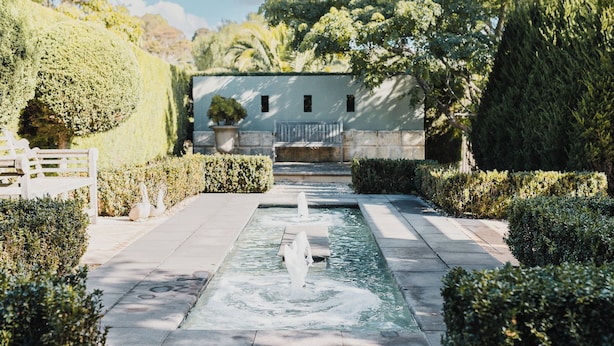 A garden pathway leading to a building with a fountain, surrounded by neatly trimmed hedges and potted plants. - Studio 22: Garden Serenity & Coastal Calm (Port Lincoln)