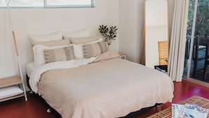 A bed with a beige blanket, a bedside table with a plant, a large mirror, and a window with curtains. - Studio 22: Garden Serenity & Coastal Calm (Port Lincoln)