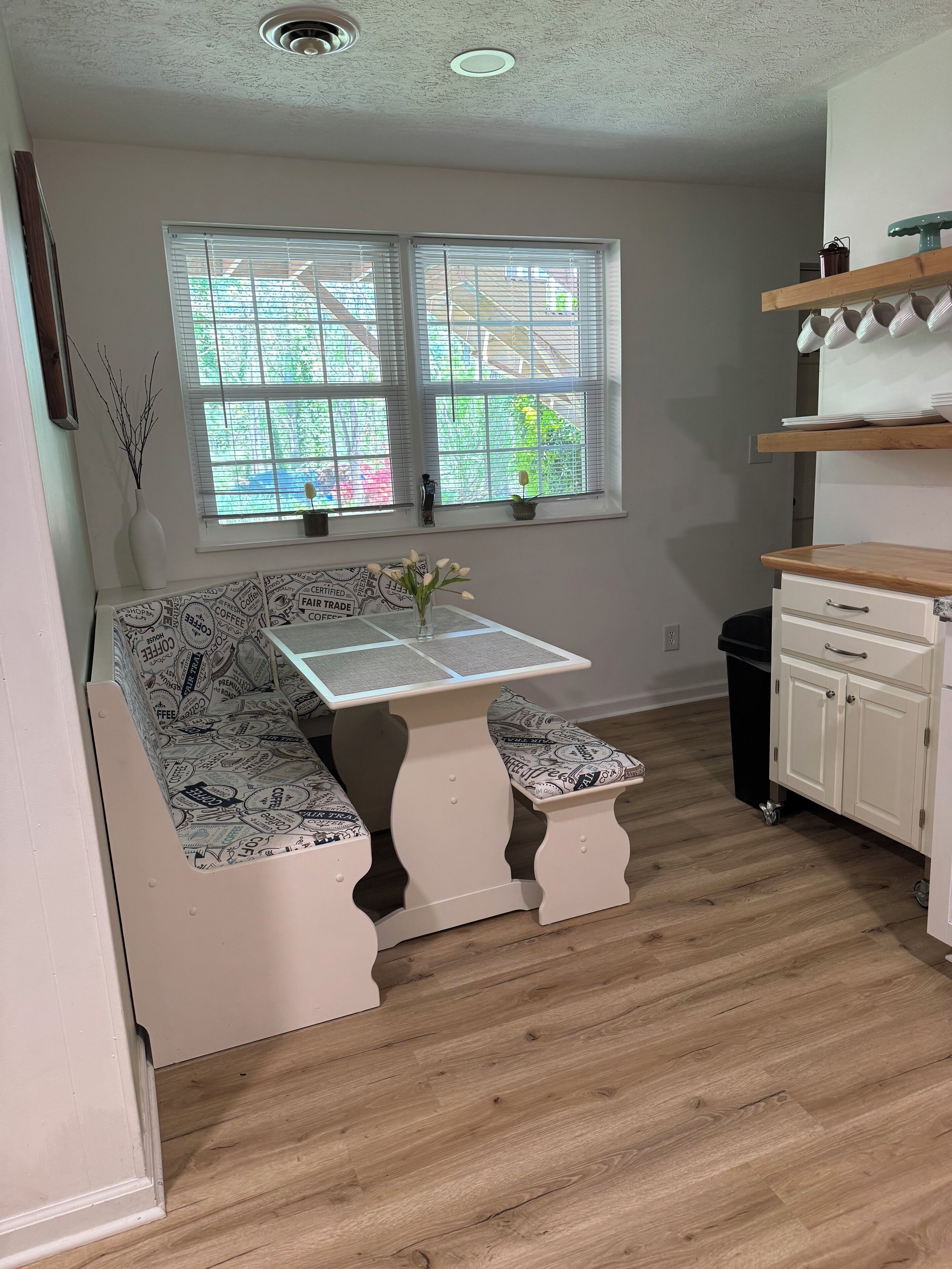 A kitchen with a wooden floor, white cabinets, a white bench with a coffee-themed table, and a window with blinds.