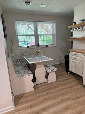 Una cocina con suelo de madera, armarios blancos, un banco blanco con una mesa con motivos de café y una ventana con persianas.