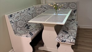Eine Sitzecke mit einer Bank und einem Tisch, ein Fenster mit Jalousien und eine Vase mit Blumen.
