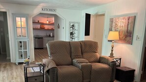 Ein Wohnzimmer mit einem beigefarbenen Ecksofa, einem gemusterten Teppich, einem Deckenventilator, einer Küche im Hintergrund und einer Tür mit der Beschriftung 'KITCHEN'.