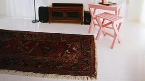 A living room with a flat-screen TV, a wooden entertainment unit, a pink table and chairs, a patterned rug, and sheer curtains. - Windy's Leisure Villa  (Negombo)