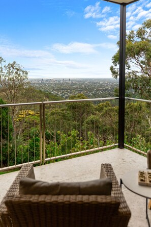 A balcony with wicker furniture and a table, overlooking a cityscape and dense greenery. - Sky Suite - A Panoramic Designer Studio in the Hills (Belair)