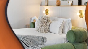 Un dormitorio con cama, almohadas, un banco tapizado en verde, un espejo y una lámpara de pared.