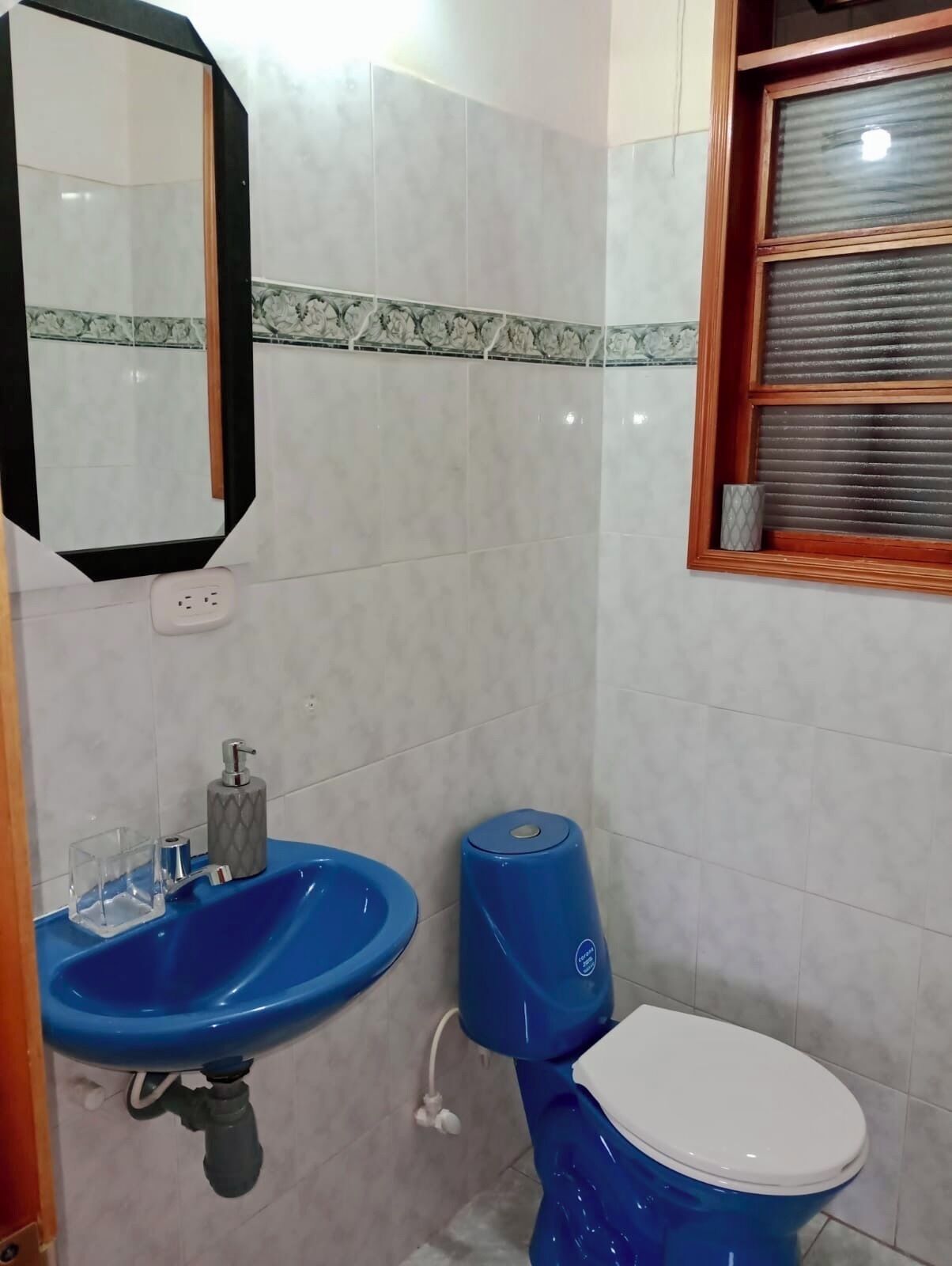 Une salle de bain avec un lavabo bleu, des toilettes, un miroir et une fenêtre avec des stores.