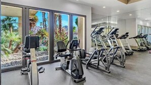 Un gimnasio con equipo de ejercicio, ventanas amplias con vistas a palmeras y un diseño interior moderno.