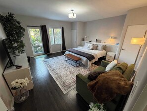 A bedroom with a bed, bedside tables, a bench, a green sofa, a floor lamp, a potted plant, and a view of the outdoors.