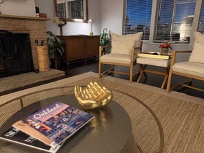 A living room with a fireplace, two chairs, a coffee table with a magazine, and a window with blinds.