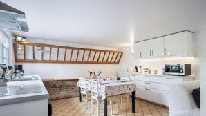 Una cocina compacta con armarios blancos, techo con vigas de madera, una mesa de comedor con sillas y un televisor.