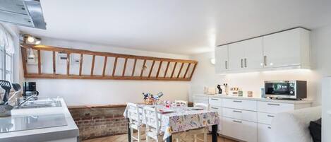 Una cocina compacta con armarios blancos, techo con vigas de madera, una mesa de comedor con sillas y un televisor.