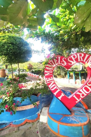 Sydämenmuotoinen kyltti, jossa lukee sanat »LOVE SPRING» ja »LOVE US», on sijoitettu puutarhaympäristöön, jossa on sinisiä kukkaruukkuja ja vihreää lehtokasvillisuutta.